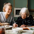 Seniorin und Pflegekraft sitzen am Tisch, beide Lächeln