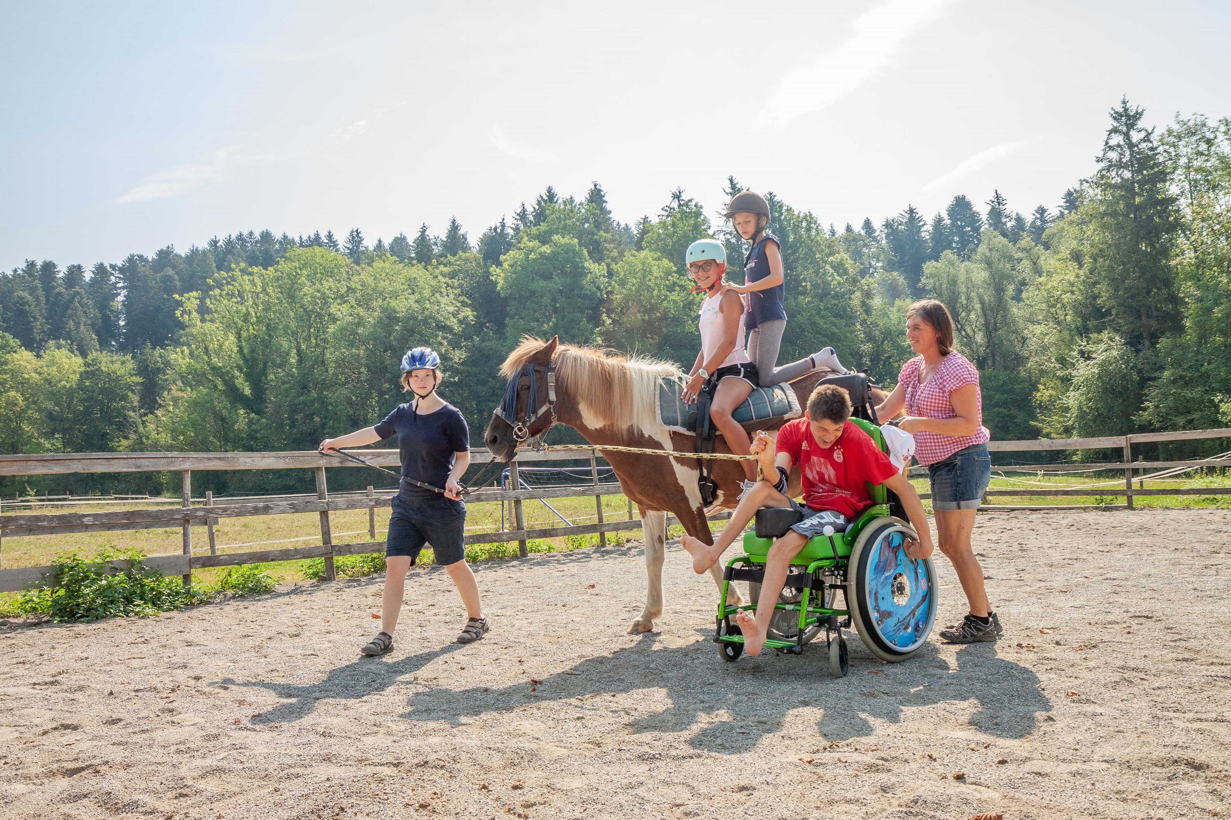 Eine Jugendliche führt ein Pferd, auf dem zwei weitere Mädchen sitzen. Neben ihnen schiebt eine Frau einen Jungen im Rollstuhl.