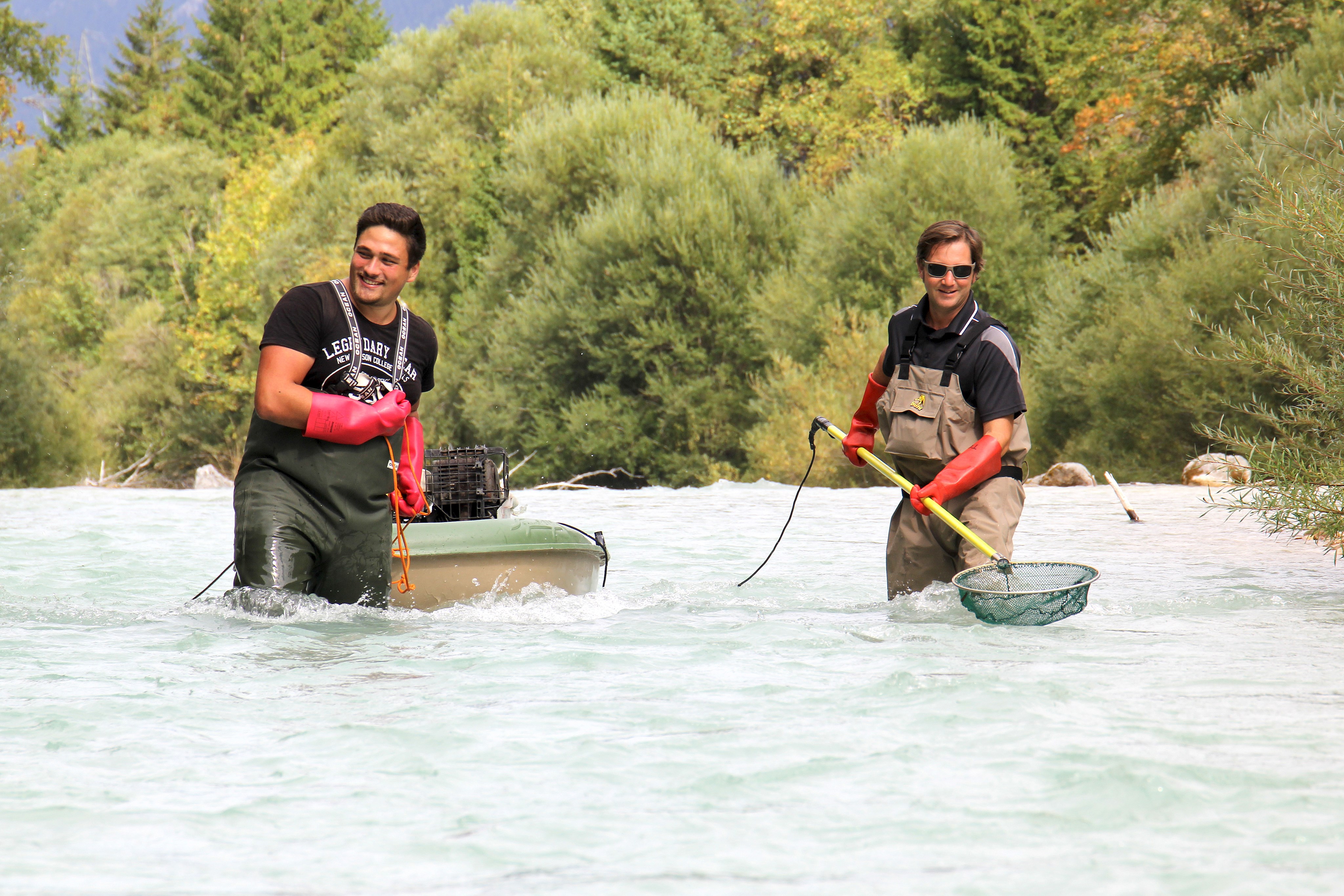 Zwei Männer stehen bis zum Oberschenkel in einem Fluss. Einer zieht ein Boot, der andere hat einen speziellen Kescher in den Händen.