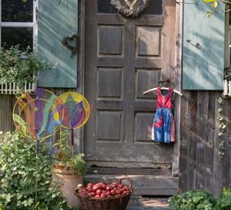Ein Kindermieder hängt vor einer alten Holztüre und ein Korb roter Äpfel stehen draußen umringt von mehreren Grünpflanzen